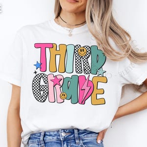 May include: White t-shirt with the words "THIRD GRADE" in colorful, retro-style lettering. The design includes stars, a lightning bolt, and smiley faces. The shirt is a casual, everyday wear item.