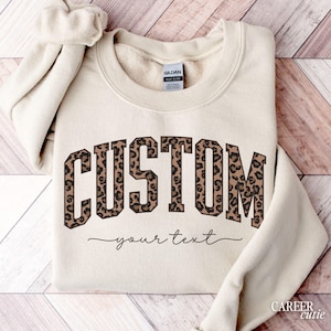 May include: A cream-colored sweatshirt with the word "CUSTOM" in a leopard print design. Below the word "CUSTOM" is the text "your text" in a cursive font. The sweatshirt is folded on a wooden surface.