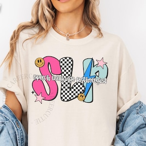 May include: Ivory-colored t-shirt with a colorful graphic design. The design features the letters "SLP" in pink, black and white checkered, and light blue, with the words "speech language pathologist" below. Smiley faces and stars are also included.