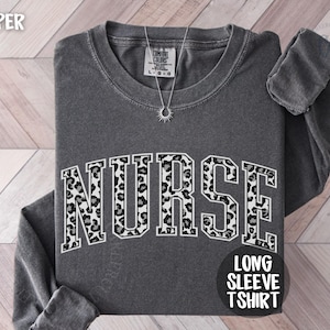 May include: A dark gray long-sleeve t-shirt with the word "NURSE" in large, bold letters across the front. The letters have a leopard print design. A silver necklace with a sun pendant is on the shirt. The words "LONG SLEEVE TSHIRT" are printed on a black circle.
