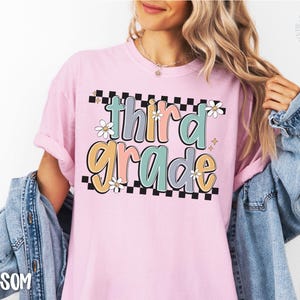 Op de afbeelding: Lichtroze T-shirt met de woorden "third grade" in een kleurrijk, retro lettertype. De tekst is zwart omlijnd en geplaatst tegen een zwart-witte geblokte achtergrond, met madeliefjes accenten.
