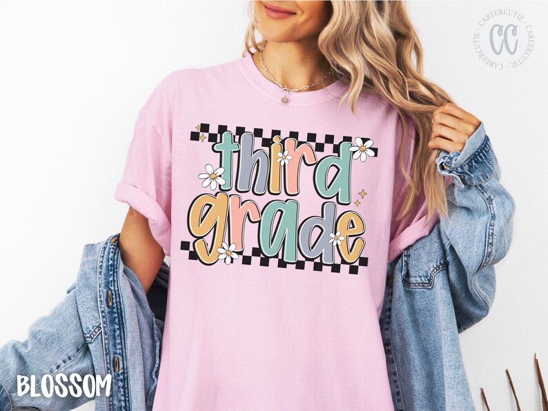 Op de afbeelding: Lichtroze T-shirt met de woorden "third grade" in een kleurrijk, retro lettertype. De tekst is zwart omlijnd en geplaatst tegen een zwart-witte geblokte achtergrond, met madeliefjes accenten.