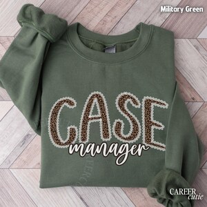 May include: Military green sweatshirt with the words "CASE manager" in a leopard print design with a white lace trim. The sweatshirt has long sleeves and is made of a soft material. The text "CAREER cutie" is in the bottom right corner.
