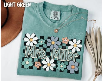 May include: Light green t-shirt with a floral design and the text "Mrs. Miller" in a retro font. The shirt features a checkerboard pattern and various flower illustrations in white, blue, and peach colors. A silver necklace with a pendant is also visible.