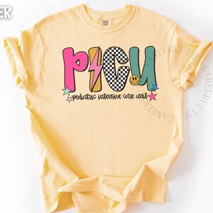 May include: Butter-colored t-shirt with the text "PICU" in colorful, playful letters. The design includes a lightning bolt, a checkerboard pattern, and a smiling face. Below the letters, it reads "pediatric intensive care unit."