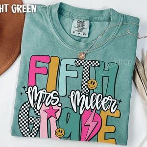 May include: Light green t-shirt with the words "FIFTH Mrs. Miller GRADE" in colorful, retro-style lettering. The design includes a checkered pattern, smiley faces, and a pink star and lightning bolt. A silver necklace with a pendant is on the shirt.