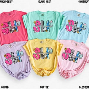 May include: A collection of six short-sleeved t-shirts in various colors, including pink, light blue, yellow, and lavender. Each shirt features a graphic design with the letters "SH" in a playful font, accompanied by stars and a checkerboard pattern.