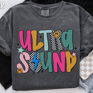 May include: Dark gray sweatshirt with the words "ULTRA SOUND" in colorful, retro-style lettering. The design includes a smiley face, a lightning bolt, and stars. The sweatshirt is displayed on a denim background.