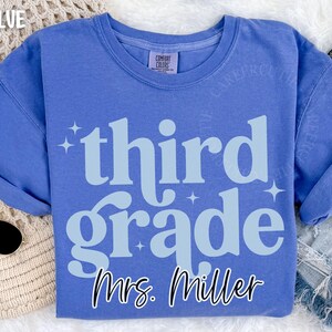 May include: Flo Blue t-shirt with the words "third grade" in light blue, with Mrs. Miller's name below. The shirt has a crew neck and short sleeves. The shirt is made of a soft material.