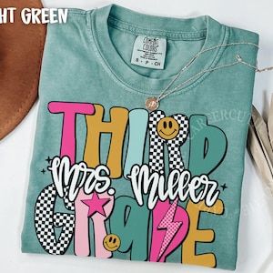 May include: Light green t-shirt with the words "THIRD Mrs. Miller GRADE" in a colorful, retro design. The design includes a smiley face, a star, and a lightning bolt. A silver necklace with a pendant is on the shirt.