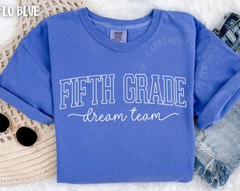 May include: A periwinkle blue short-sleeve t-shirt featuring the white text FIFTH GRADE in a collegiate block font, followed by the word dream team in a white cursive script. The shirt has a crew neckline and a visible brand label at the neck.