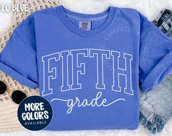Puede incluir: Una camiseta azul Flo con las palabras "FIFTH grade" en letras blancas y cursiva. La camiseta tiene cuello redondo y mangas cortas. Un círculo azul marino con las palabras "MORE COLORS AVAILABLE" está en la esquina inferior izquierda.