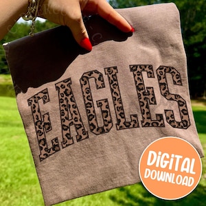 Pode incluir: Uma camiseta marrom com a palavra "EAGLES" em letras grandes com estampa de leopardo. A camiseta est&aacute; sendo segurada por uma m&atilde;o com esmalte vermelho. Um c&iacute;rculo laranja com as palavras "DOWNLOAD DIGITAL" est&aacute; no canto inferior direito.