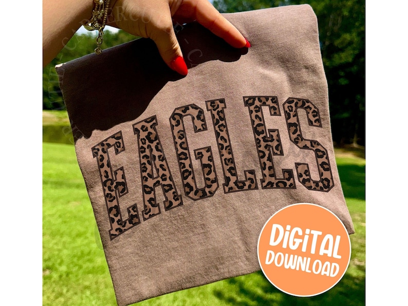 Pode incluir: Uma camiseta marrom com a palavra "EAGLES" em letras grandes com estampa de leopardo. A camiseta est&aacute; sendo segurada por uma m&atilde;o com esmalte vermelho. Um c&iacute;rculo laranja com as palavras "DOWNLOAD DIGITAL" est&aacute; no canto inferior direito.