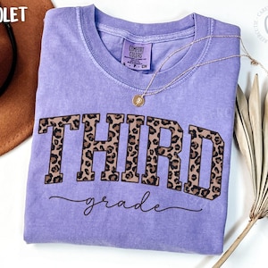May include: A violet Comfort Colors t-shirt with the words "THIRD grade" in a leopard print design. A gold necklace with a pendant is on the shirt. A brown hat and a dried palm leaf are in the background.