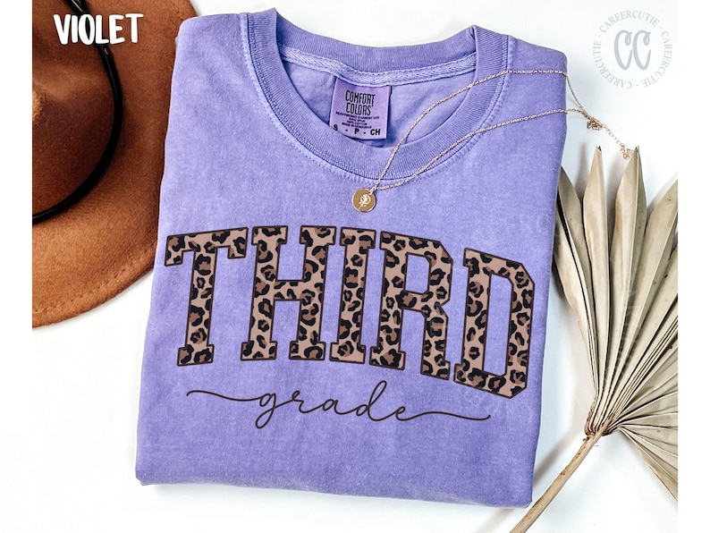 May include: A violet Comfort Colors t-shirt with the words "THIRD grade" in a leopard print design. A gold necklace with a pendant is on the shirt. A brown hat and a dried palm leaf are in the background.