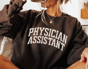 May include: A dark chocolate-coloured sweatshirt featuring the words "PHYSICIAN ASSISTANT" in white, block lettering. The crew neck sweatshirt is made of a soft material. A person is holding a black mug in the image.