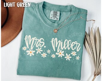 May include: Light green t-shirt with the text "Mrs. Miller" in a cursive font, decorated with white daisy flowers. A silver necklace with a pendant rests on the shirt. The shirt is a Comfort Colors brand.