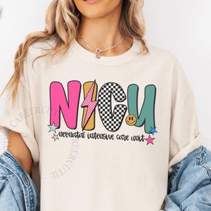 May include: Ivory-colored t-shirt with a colorful NICU graphic. The design features the letters "NICU" in pink, black and white checkered, and teal, with a lightning bolt and smiley face. The words "neonatal intensive care unit" are below.