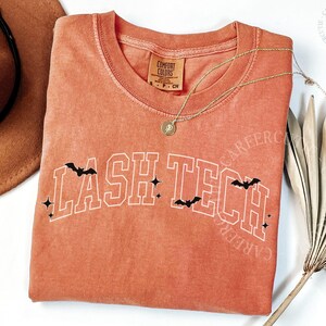 May include: An orange-red t-shirt with the text "LASH TECH" in white outlined letters, decorated with black bats and stars. A gold necklace with a pendant is draped across the shirt. A brown hat is visible in the background.