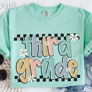 Op de afbeelding: Een mintgroen t-shirt met de woorden "third grade" in een retro lettertype. De tekst is in pastelkleuren met madeliefjesaccenten en een zwart-wit geblokte rand. Zonnebril en armbanden zijn zichtbaar.
