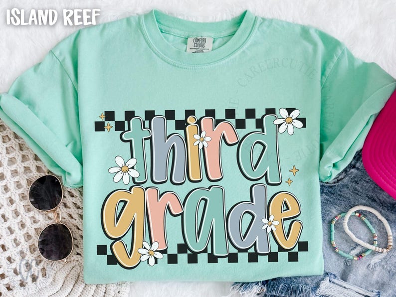 Op de afbeelding: Een mintgroen t-shirt met de woorden "third grade" in een retro lettertype. De tekst is in pastelkleuren met madeliefjesaccenten en een zwart-wit geblokte rand. Zonnebril en armbanden zijn zichtbaar.