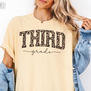 May include: Butter-colored t-shirt with the words "THIRD grade" in a bold, leopard print font. The word "grade" is in a cursive font. The shirt has rolled-up sleeves and is paired with a denim jacket.