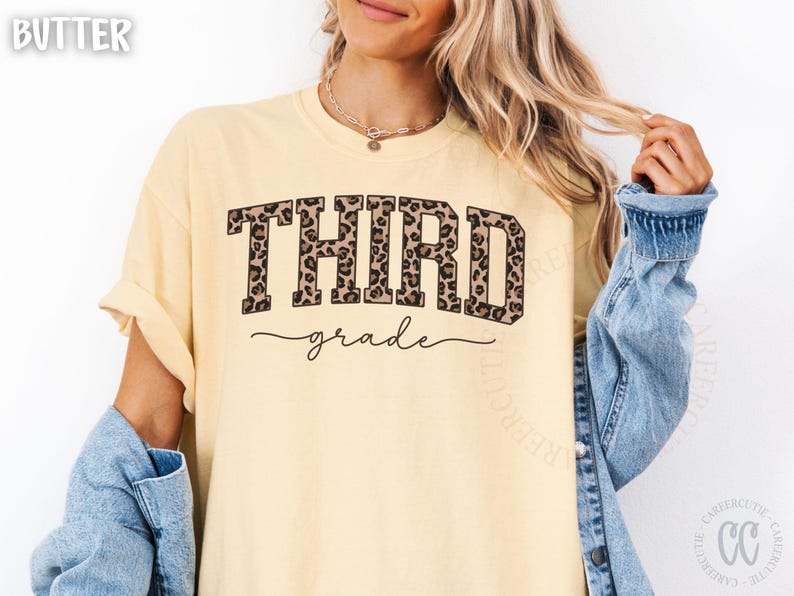 May include: Butter-colored t-shirt with the words "THIRD grade" in a bold, leopard print font. The word "grade" is in a cursive font. The shirt has rolled-up sleeves and is paired with a denim jacket.