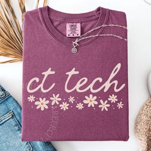 May include: A maroon t-shirt with the words "ct tech" in a cursive font, decorated with daisy graphics. A silver necklace and a pair of jeans are next to the shirt. The shirt is from Comfort Colors.
