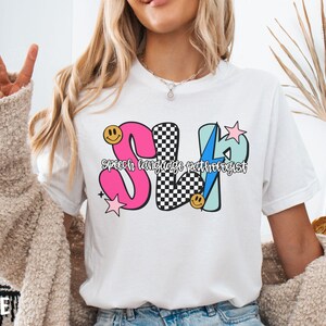 May include: White t-shirt with a colorful graphic design. The design features the letters "SLP" in pink, black and white checkered, and blue, with the words "Speech language pathologist" underneath. Smiley faces and stars are also included.