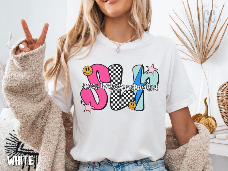 May include: White t-shirt with a colorful graphic design. The design features the letters "SLP" in pink, black and white checkered, and blue, with the words "Speech language pathologist" underneath. Smiley faces and stars are also included.