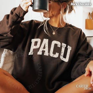 May include: Dark chocolate brown sweatshirt with the word "PACU" in distressed white lettering. The sweatshirt has a crew neck and long sleeves. The text "CAREER cutie" is printed on the bottom right corner of the sweatshirt.