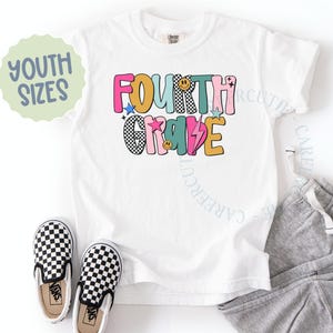 K&ouml;nnte beinhalten: Wei&szlig;es T-Shirt mit den Worten "FOURTH GRADE" in farbenfrohen, verspielten Buchstaben. Das Design umfasst Sterne, einen Blitz und ein Smiley. Ein Paar schwarz-wei&szlig; karierte Slip-On-Schuhe und eine graue Jogginghose sind ebenfalls im Bild.