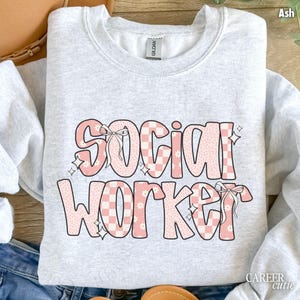 May include: Ash-colored sweatshirt with the words "SOCIAL WORKER" in a pink, checkered, and floral design. The text includes bows and star accents. The sweatshirt is a crew neck style.