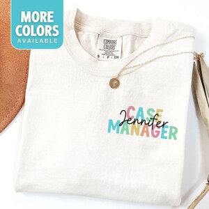 May include: A white t-shirt with the text "CASE MANAGER" in rainbow colors. The name "Jennifer" is written in black cursive script above the text. The shirt is folded and lying on a brown surface.