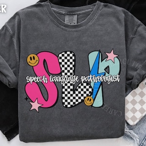 May include: Dark gray t-shirt with a colorful graphic design. The design features the letters "SLP" in pink, checkered, and light blue, with the words "speech language pathologist" in white. Smiley faces and stars accent the design.