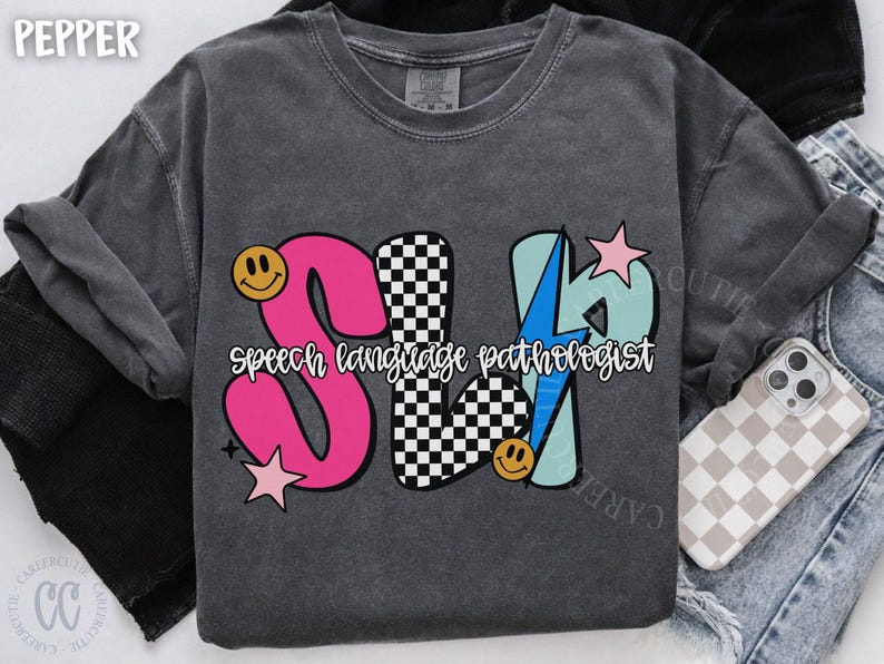 May include: Dark gray t-shirt with a colorful graphic design. The design features the letters "SLP" in pink, checkered, and light blue, with the words "speech language pathologist" in white. Smiley faces and stars accent the design.