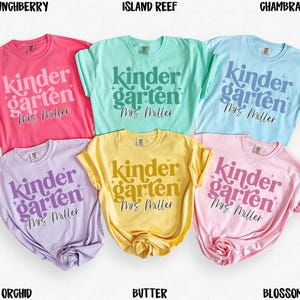 May include: A collection of six short-sleeved t-shirts in various colors, including pink, green, blue, purple, yellow, and light pink. Each shirt features the words "Kindergarten Mrs. Miller" in a decorative font. The shirts are likely for teachers or students.