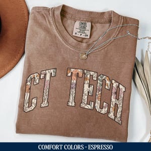 May include: A brown t-shirt with a floral design spelling out "CT TECH" in white letters. The shirt is made by Comfort Colors and is in the color Espresso.