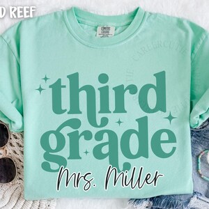 May include: A pale green t-shirt with the words "third grade" in teal, and the name "Mrs. Miller" below. The shirt has a crew neck and short sleeves. Sunglasses and bracelets are visible.