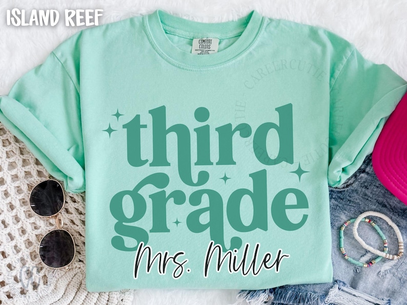 May include: A pale green t-shirt with the words "third grade" in teal, and the name "Mrs. Miller" below. The shirt has a crew neck and short sleeves. Sunglasses and bracelets are visible.