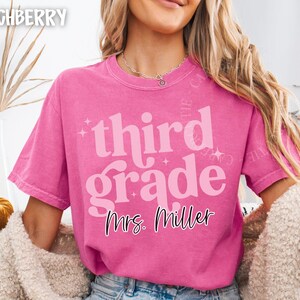 May include: A vibrant pink t-shirt featuring the text "third grade" in a retro style, with "Mrs. Miller" underneath. The shirt has a loose fit and is probably made of soft cotton. This is a teacher-themed item.