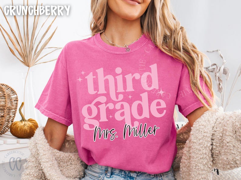 May include: A vibrant pink t-shirt featuring the text "third grade" in a retro style, with "Mrs. Miller" underneath. The shirt has a loose fit and is probably made of soft cotton. This is a teacher-themed item.