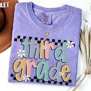 Op de afbeelding: Een violet Comfort Colors T-shirt met de woorden "third grade" in een kleurrijk, retro lettertype. Het ontwerp bevat madeliefjes en een dambordpatroon. Een gouden ketting en een bruine hoed zijn zichtbaar.