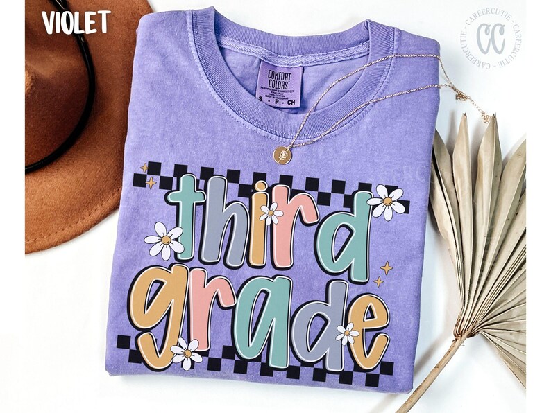 Op de afbeelding: Een violet Comfort Colors T-shirt met de woorden "third grade" in een kleurrijk, retro lettertype. Het ontwerp bevat madeliefjes en een dambordpatroon. Een gouden ketting en een bruine hoed zijn zichtbaar.