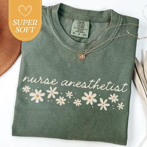 May include: A sage green t-shirt with the words "nurse anesthetist" in a cursive font, decorated with white daisy flowers. A gold necklace with a small pendant is on the shirt. The shirt has a "Super Soft" tag.