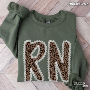 May include: Military green sweatshirt with the letters "RN" in a leopard print design, outlined with white lace trim. The sleeves are cuffed. The text "CAREER cutie" is in the lower right corner.