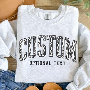 May include: A light gray sweatshirt with the word "CUSTOM" in a black zebra print font, and "OPTIONAL TEXT" below. The sweatshirt is folded on top of blue jeans.