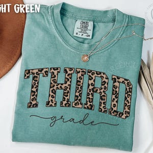 May include: Light green Comfort Colors t-shirt with "THIRD grade" in leopard print lettering. A silver necklace with a pendant is on the shirt. The shirt is a crew neck and has a tag that says "COMFORT COLORS".