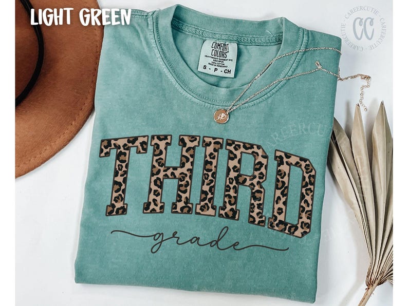 May include: Light green Comfort Colors t-shirt with "THIRD grade" in leopard print lettering. A silver necklace with a pendant is on the shirt. The shirt is a crew neck and has a tag that says "COMFORT COLORS".